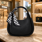 A black pebbled leather hobo handbag with a top handle, featuring a black and white checkered scarf tied to the handle, displayed on a marble counter in a luxury boutique.