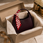 A burgundy pebbled leather handbag with a red and white checkered silk scarf tied to its handle, presented inside a beige Heritage box with branded tissue paper.