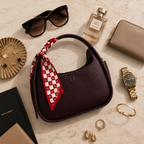 A dark burgundy pebbled leather handbag with a red and white checkered silk scarf tied to its handle, styled in a luxury flat-lay with sunglasses, a perfume bottle, a wallet, jewelry, and watches on a beige marble background.