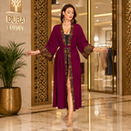 A woman stands barefoot in a luxury indoor setting wearing a long magenta-purple robe with gold and black geometric patterns along the collar, cuffs, and front opening. The background features a marble floor, decorative gold-toned columns, and a plant.