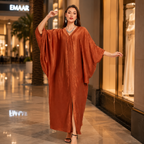 A woman modeling a flowing, rust-orange kaftan dress in a luxury shopping mall corridor. The dress features a V-neckline, wide draped sleeves, and a high front slit.