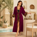 A woman stands barefoot in a sunlit patio setting wearing a long magenta-purple open-front robe with wide sleeves and black-and-gold geometric trim. The robe has a front slit and a self-tie belt.