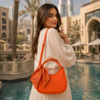 A woman in a white long-sleeve dress stands by a pool in a luxurious resort setting, wearing a bright orange handbag with a matching orange scarf accent.