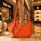 An orange pebbled leather shoulder bag with a matching orange scarf tied in a bow around the strap, displayed on a marble counter in a luxury shopping mall setting.