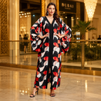 A woman models a long, flowing black maxi dress with wide sleeves and a bold abstract geometric pattern in red, white, and black, set against a luxurious indoor mall background.