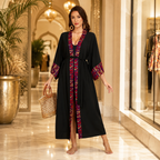 A woman stands barefoot in a luxurious indoor boutique corridor wearing a long black kaftan-style robe with ornate red, purple, and gold embroidered trim along the neckline, sleeves, and front slit. She is holding a woven straw handbag.