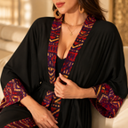 A woman wearing a black kimono-style robe with richly patterned red, purple, and gold trim along the neckline and sleeves, styled in a warm, sunlit indoor setting.